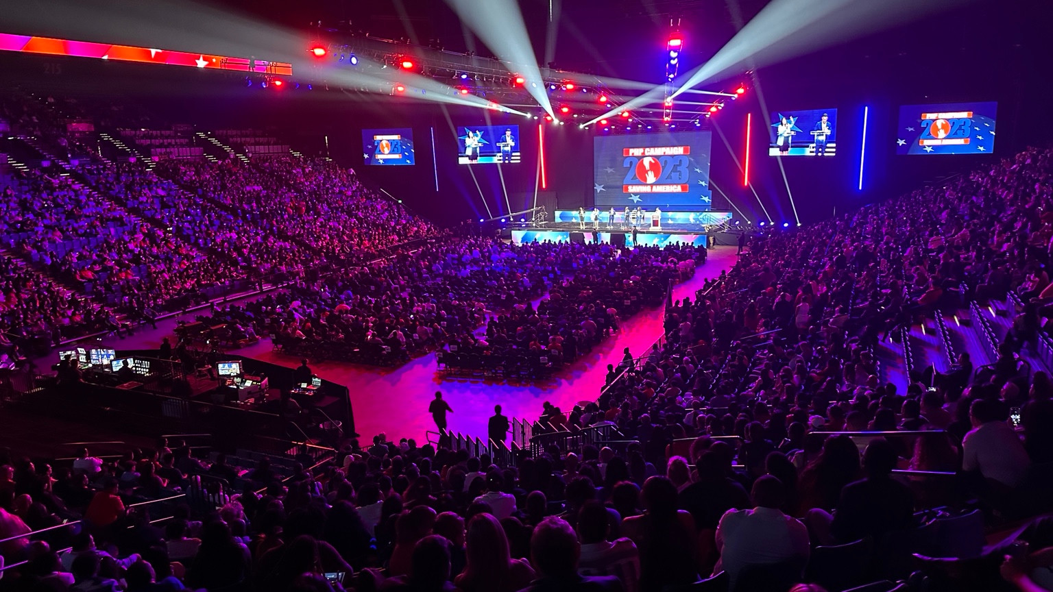 A full arena viewed from above, with a central stage, suspended lighting, large screens, and a live audience